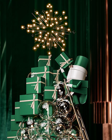 La Mer attendant caricature placing a bright, golden star at the top of stacked La Mer gift boxes in the shape of a Christmas tree